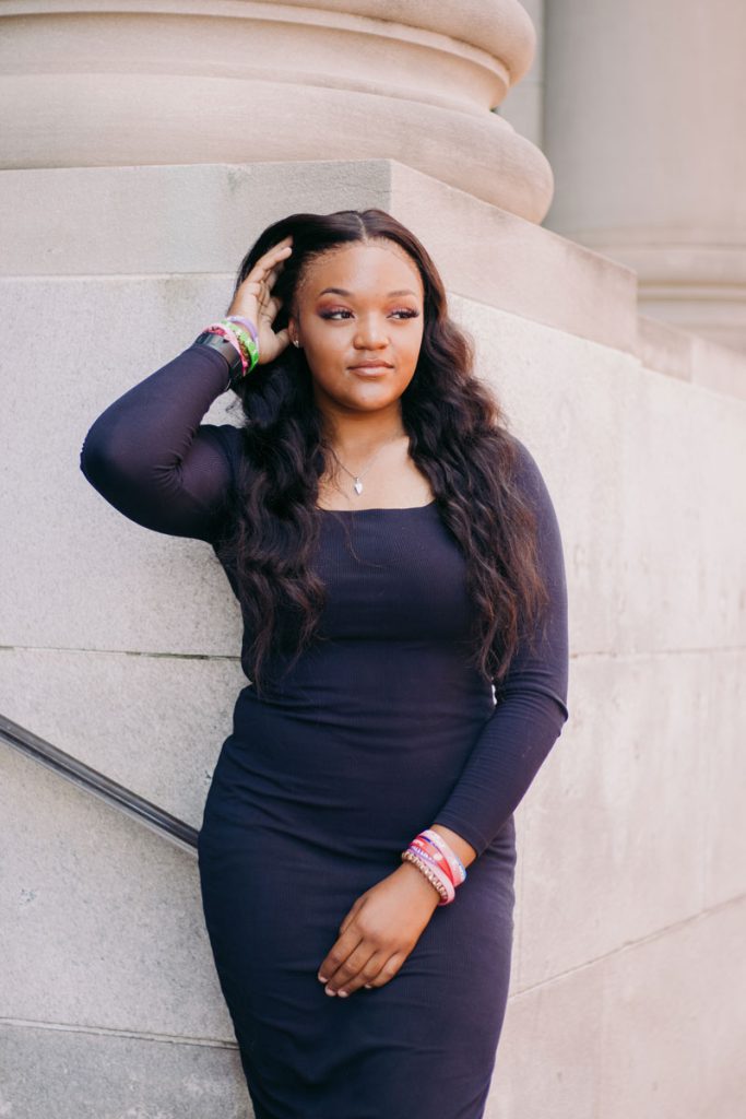 A young teen in a black dress poses outside against a marble wall