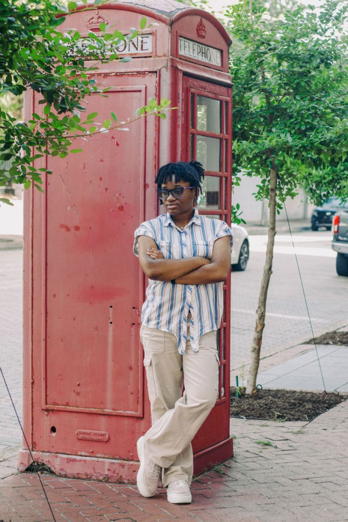 A teen in a striped shirt leans against an old red phone booth on an outside sidewalk