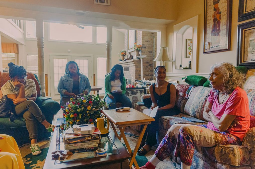 Four teenagers sit on the couch in a brightly lit living room with an older woman on the other end of the seating area