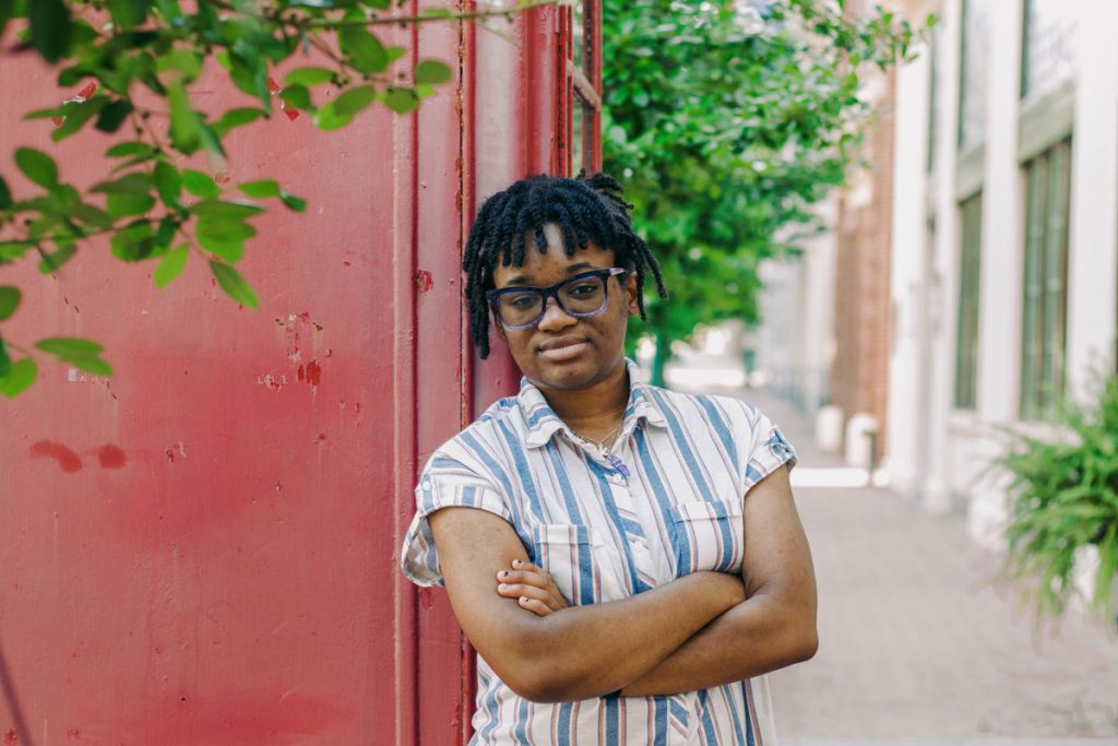 A teen in a striped shirt leans against an old red phone booth on an outside sidewalk