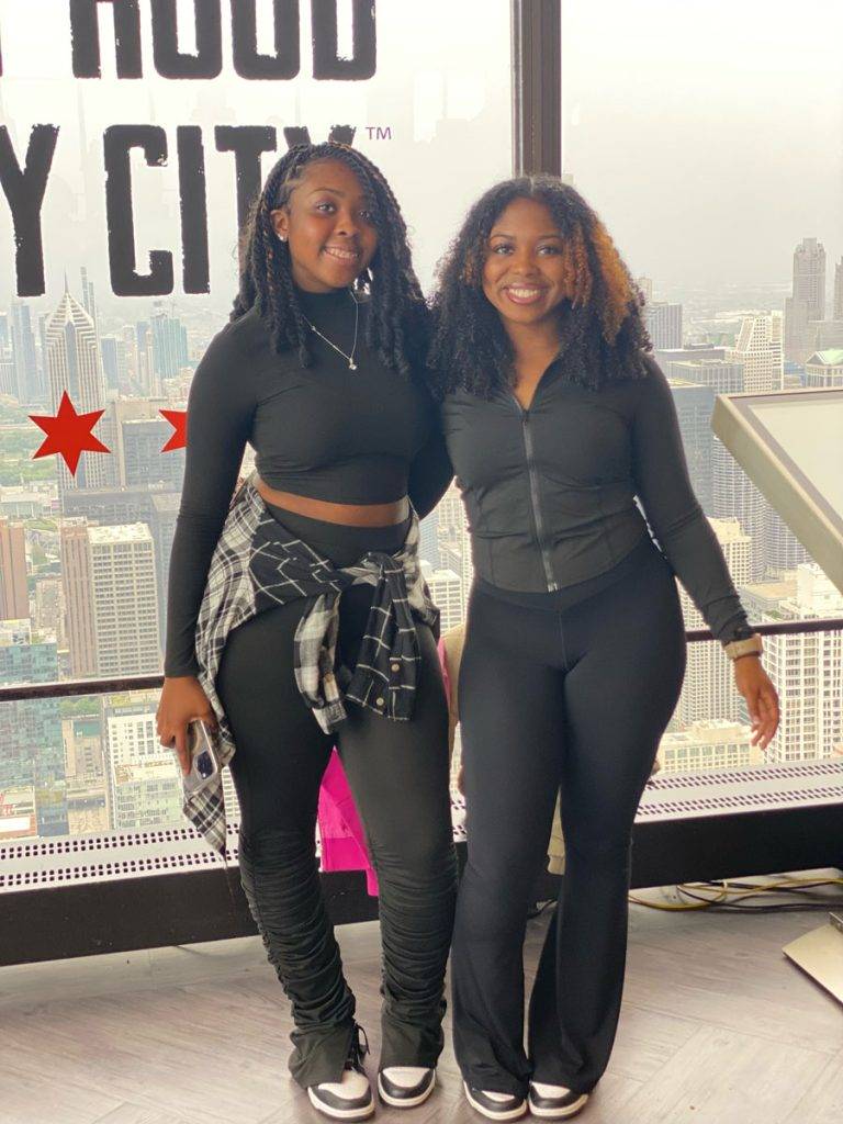 Two teens pose in front of a window with a visible city skyline