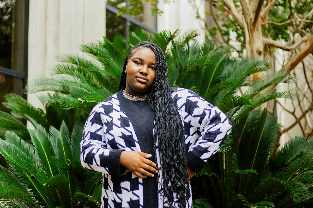 A teen with long braided hair stands outside in front of green palm fronds