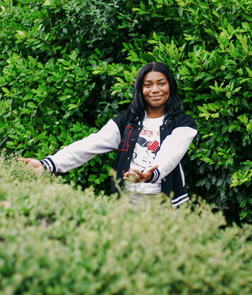 A teen stands outside in front of a wall of greenery