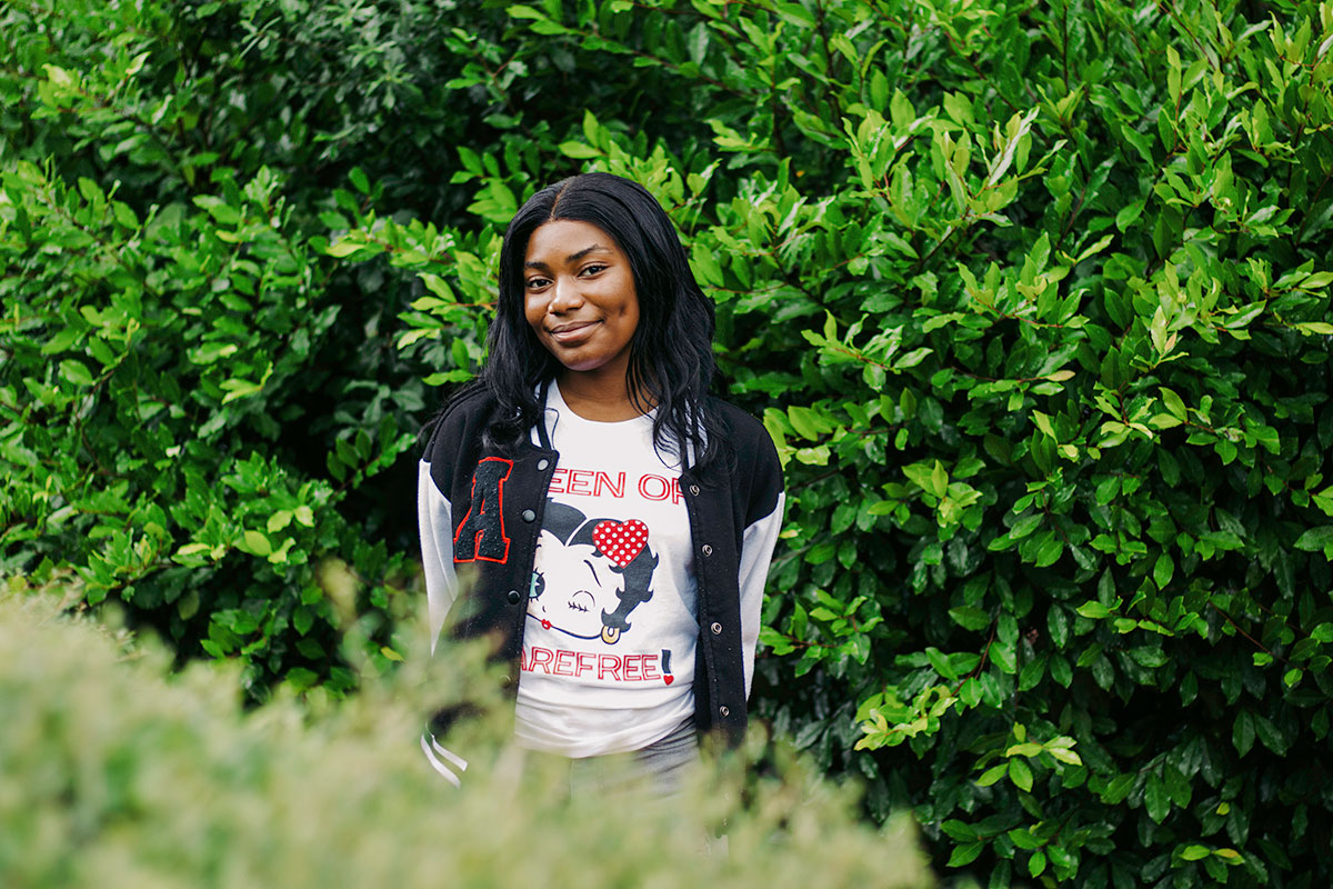 A teen stands outside in front of a wall of greenery