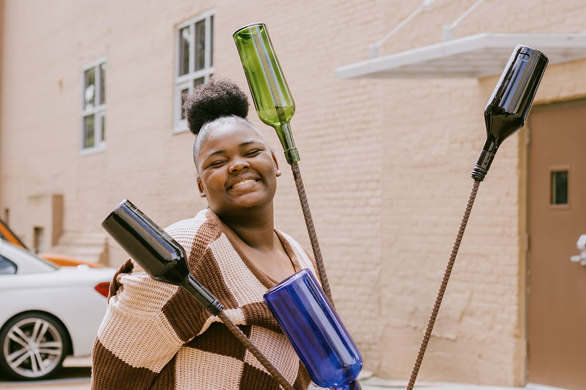 A teen in a brown checkered sweater poses outside behind a bottle tree