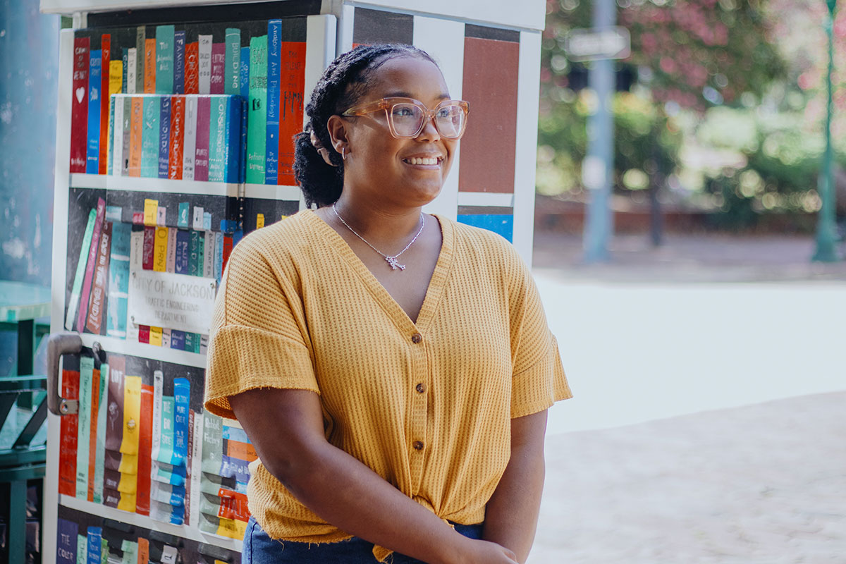 A teen in glasses and a yellow top stands outside in front of a utility box brightly painted to look like a bookshelf filled with books