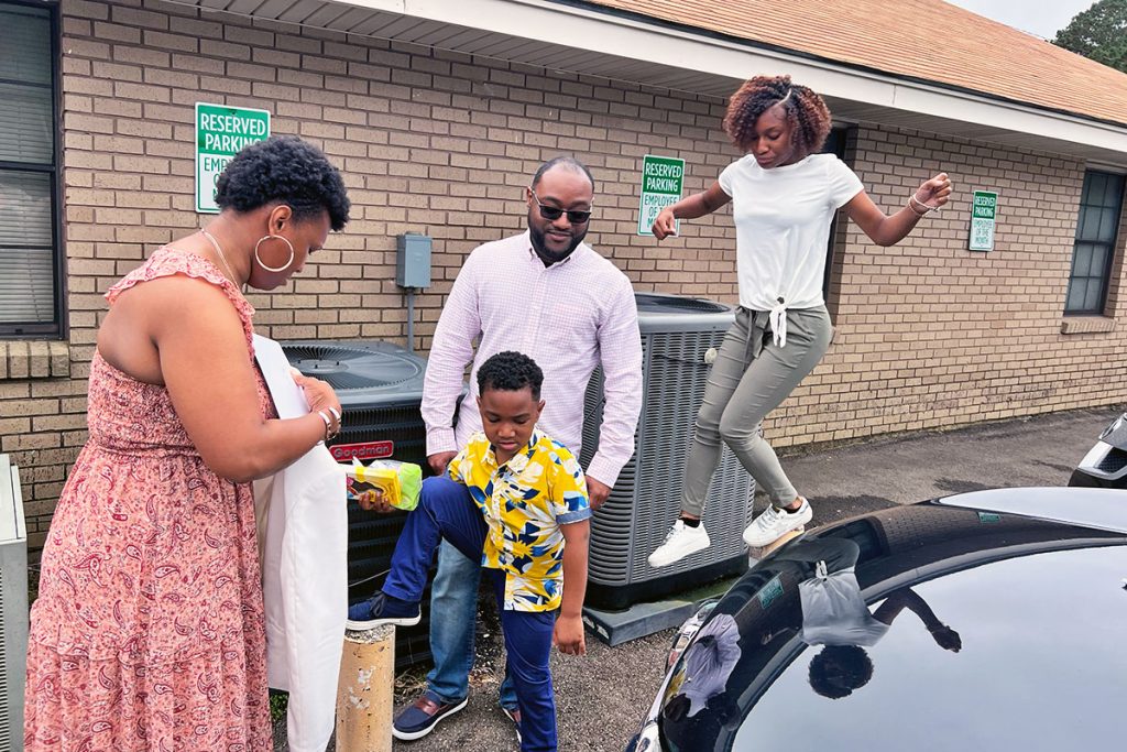 A family of four are gathered outside of a church by a car. A teen is precariously balancing on a yellow parking pole while the mother folds a piece of clothing