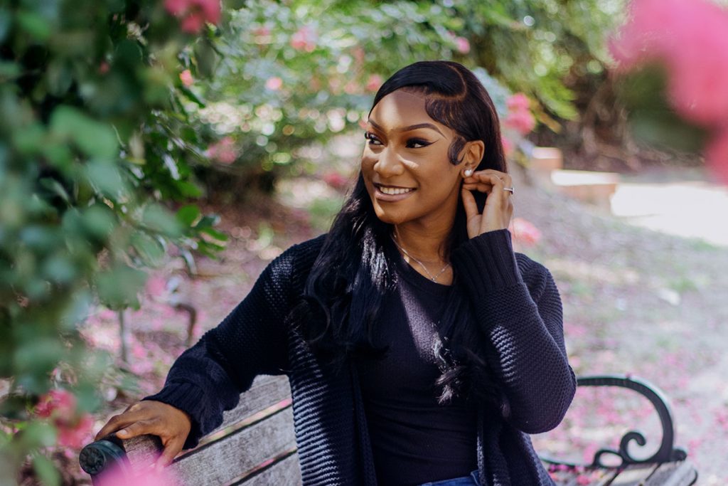 A teen poses on a bench in a park. Pink flowering bushes frame the top and left sides.