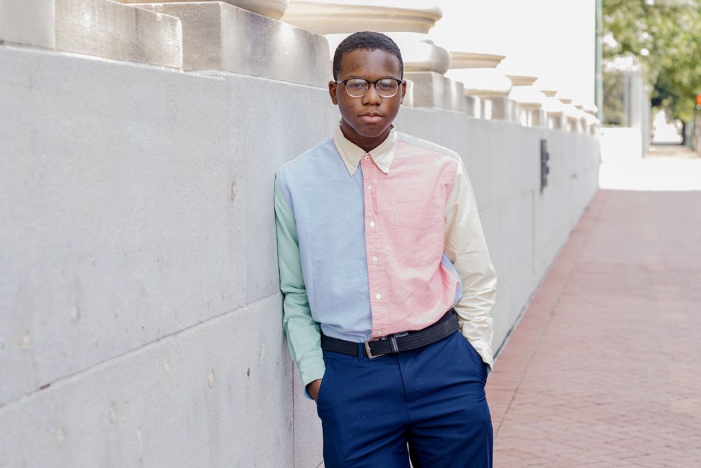 A teen poses ouside next to a building with a tall white wall. The teen is wearing a color blocked button-down shirt and blue slacks.