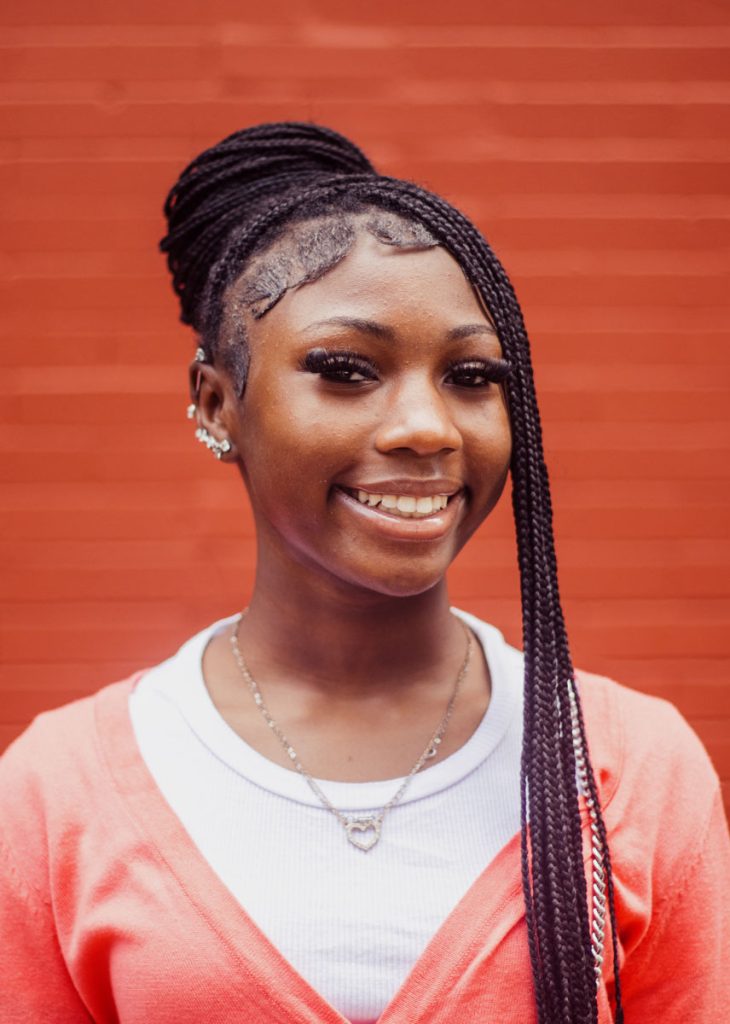 A teen with long braided hair smiles against a brick wall